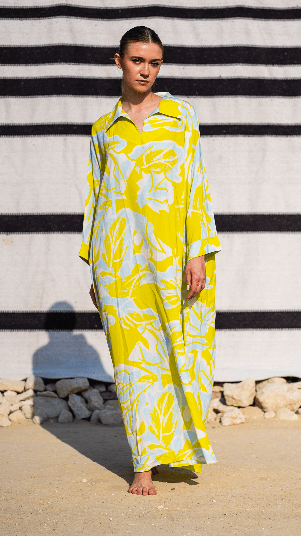 Woman wearing a yellow and white patterned dress standing against a black and white striped wall.