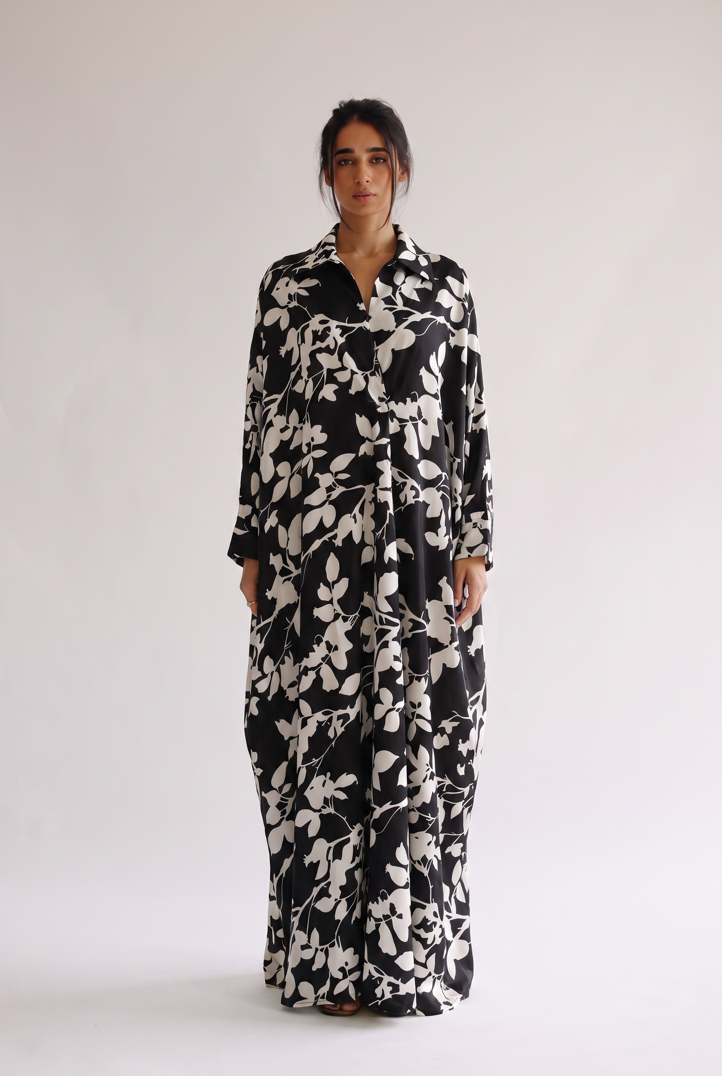 A woman modeling a long-sleeved black maxi dress with a white leafy floral pattern, standing against a plain light gray studio background.