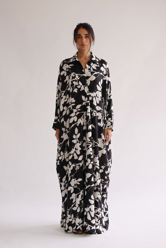 A woman modeling a long-sleeved black maxi dress with a white leafy floral pattern, standing against a plain light gray studio background.