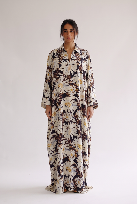 A woman stands against a plain light gray studio background wearing a long-sleeve, collared maxi dress with a large white daisy floral print over a dark brown base.